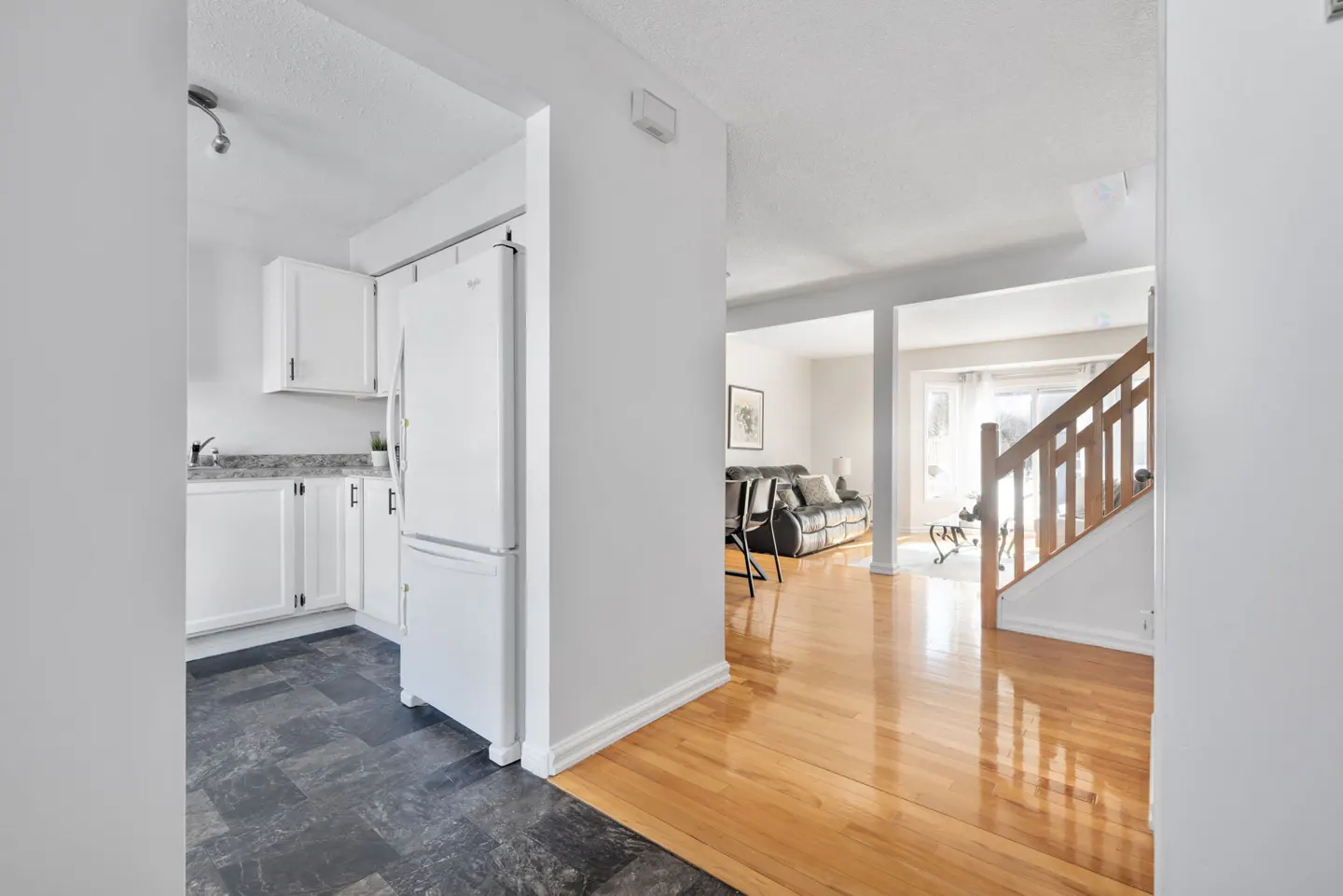 A bright, open-concept home interior with a view of the kitchen, living room, and staircase. The kitchen features white cabinets and a white refrigerator. The living room has hardwood floors and a black leather sofa.
