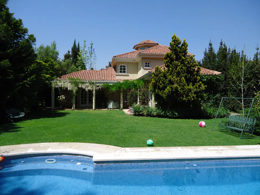 Backyard view of a tan house with a red tile roof, green lawn, pool, swing, and trees.