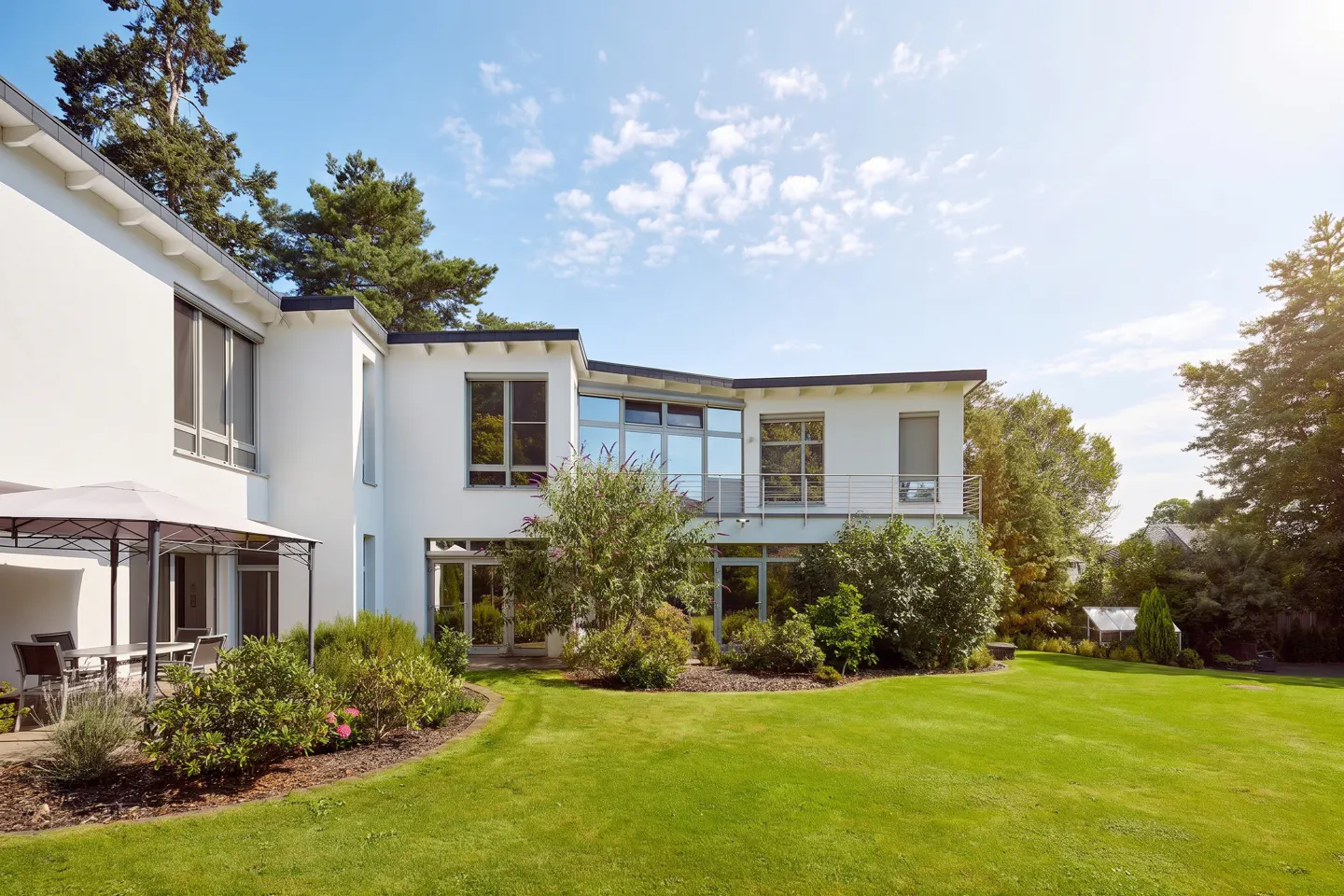 Exterior of a modern white house with large windows, green lawn, and patio furniture under a gazebo.