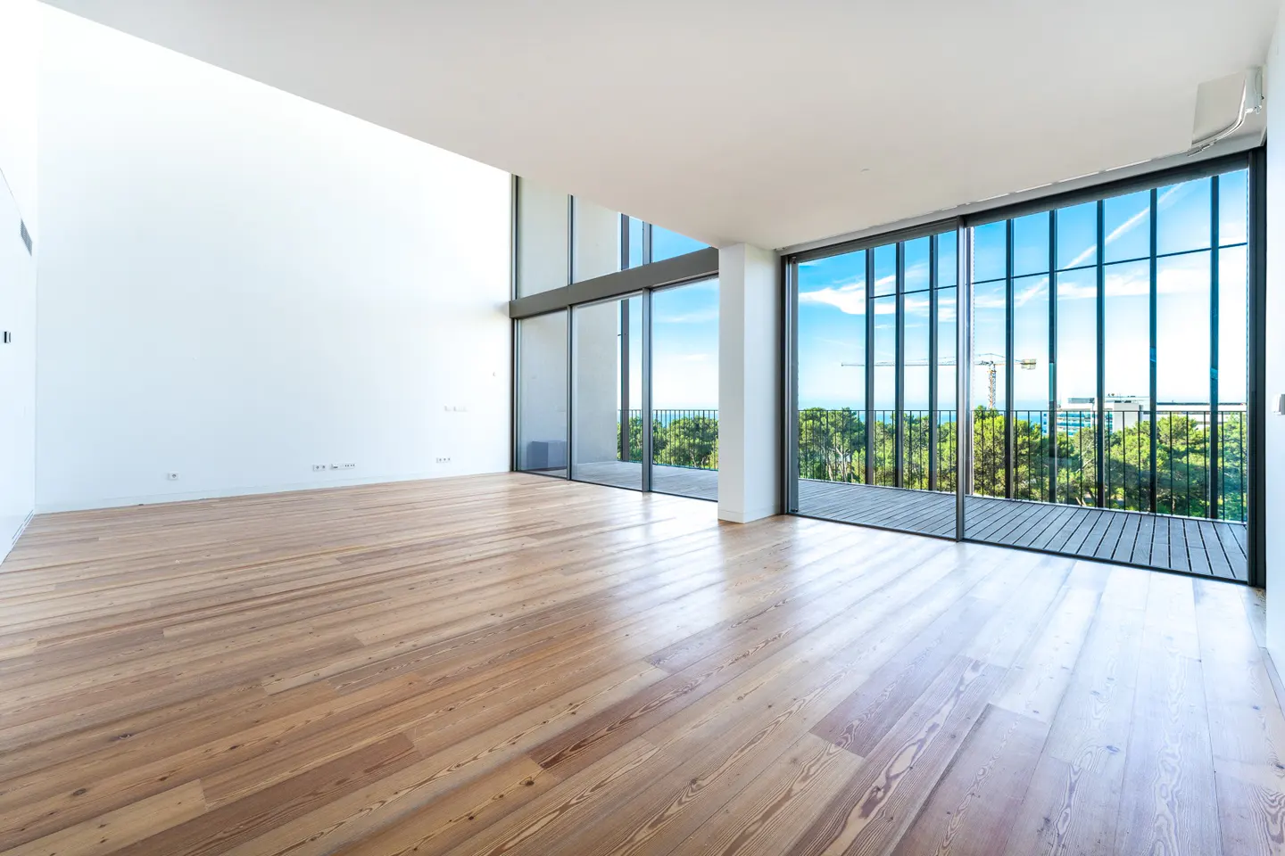 Bright, empty room with light wood floors and white walls. Large sliding glass doors open to a balcony with a view of trees and the ocean.
