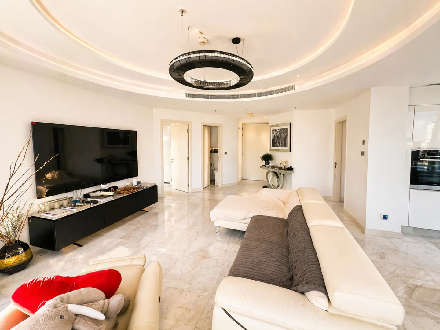 Bright, modern living room with white walls, marble floors, and a large black TV. A white sectional sofa faces the TV. A round, black chandelier hangs from the ceiling.