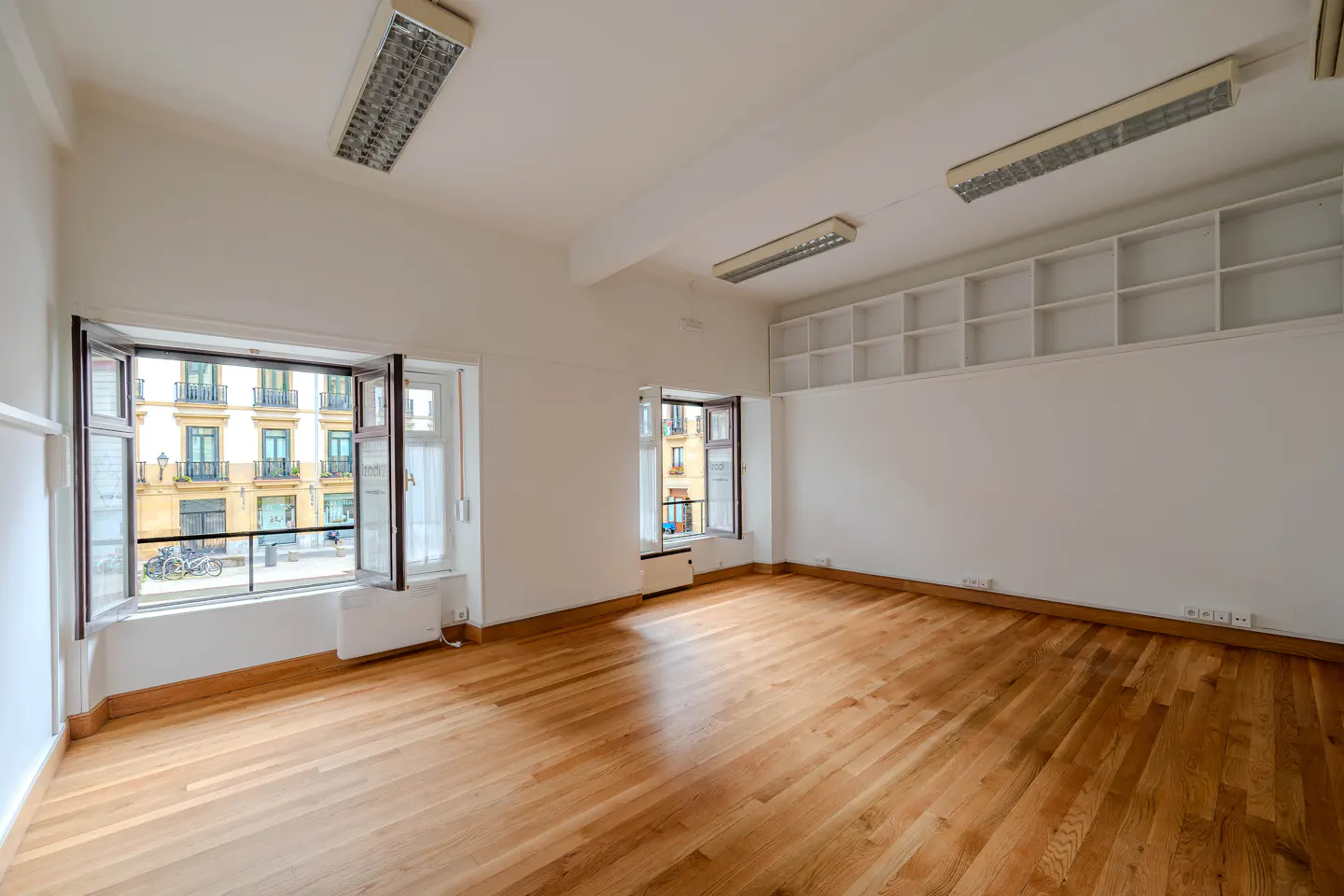 Bright, empty room with hardwood floors, white walls, and two open windows showing a city view.
