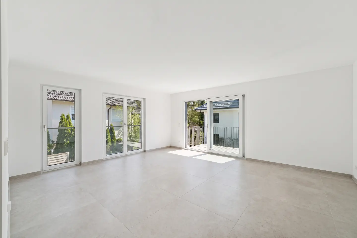 Bright, empty room with white walls and light gray tile floor. Three glass doors lead to outdoor views of greenery and a balcony.