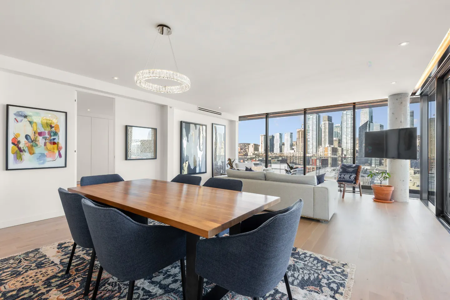 Bright, modern condo interior with a wood dining table, blue chairs, and city views from large windows. Art adorns the white walls.