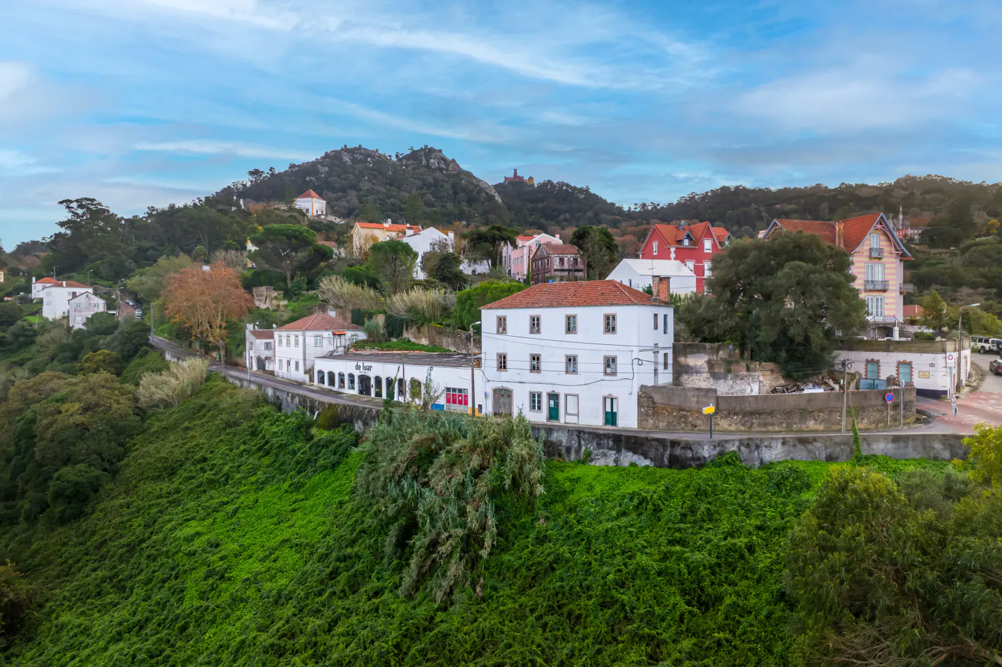 Hillside view of white buildings with red tile roofs, green foliage, and a blue sky with clouds.