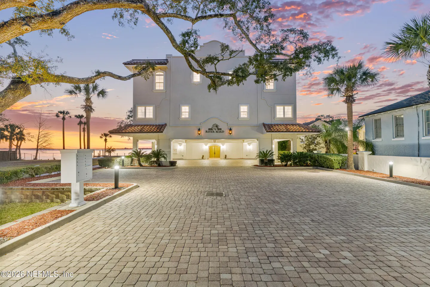 Exterior view of a white, three-story building with a brick driveway and palm trees at sunset.