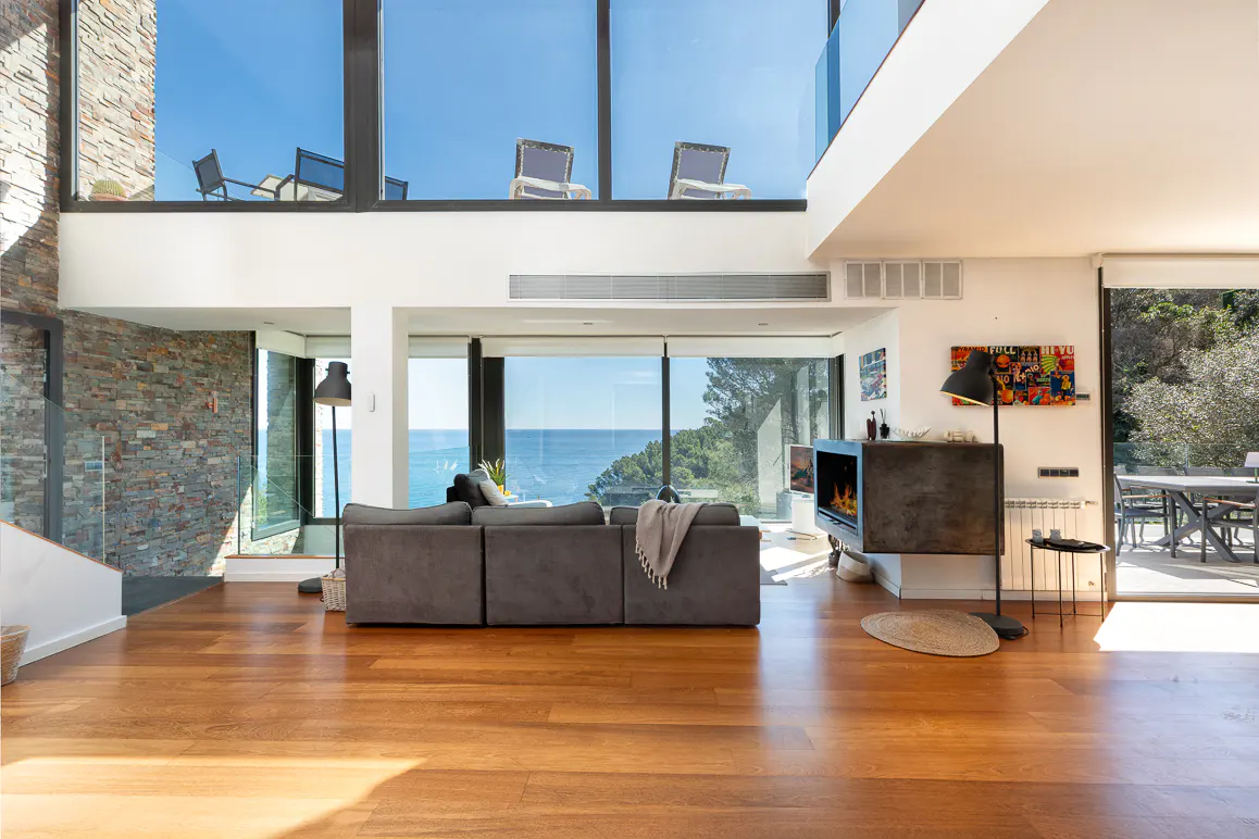 Bright, modern living room with hardwood floors, gray sofa, and ocean view through large windows. Fireplace and outdoor seating visible.