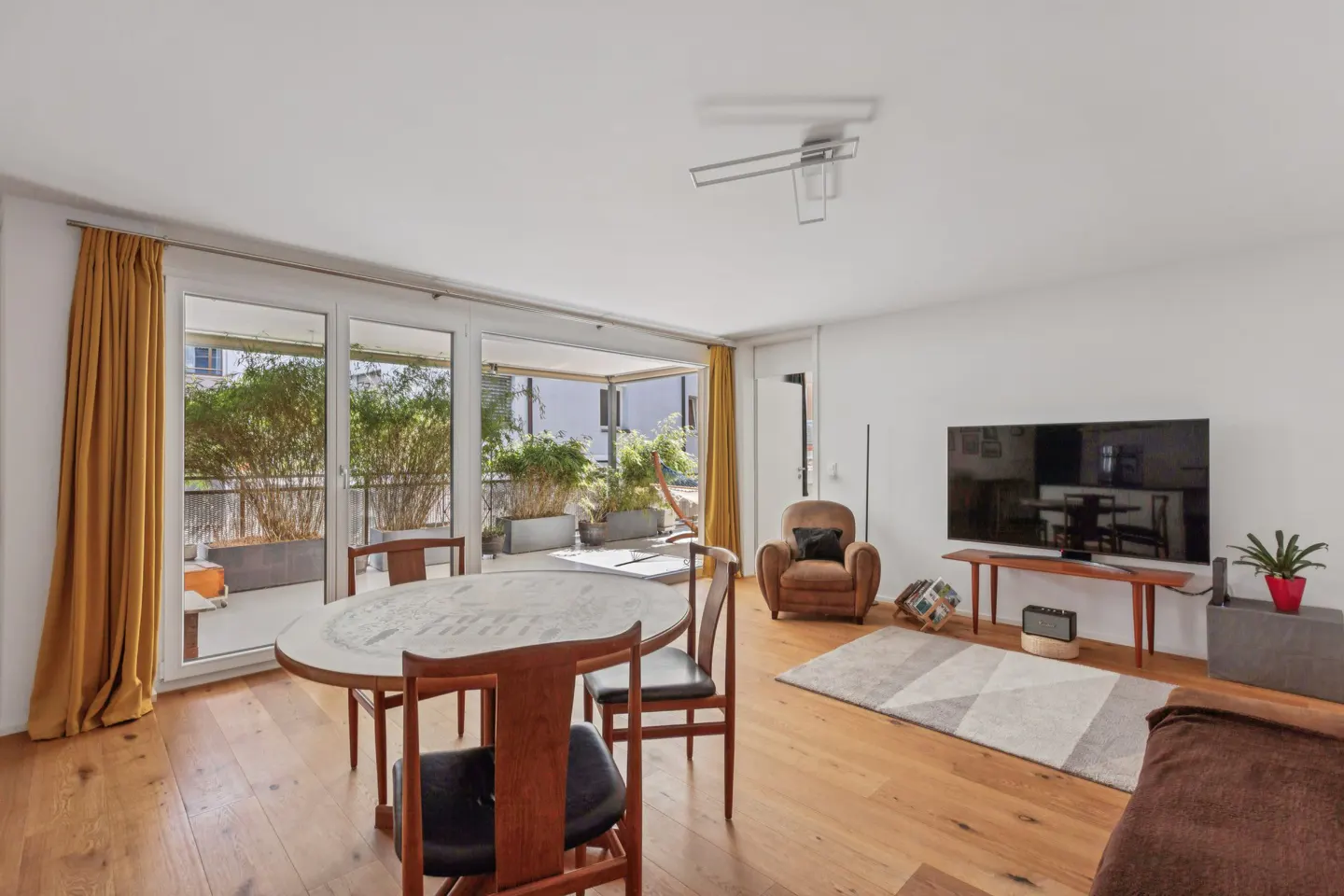 Bright living room with wood floors, a round table with chairs, and a balcony with plants visible through sliding glass doors. A brown armchair and TV are on the right.