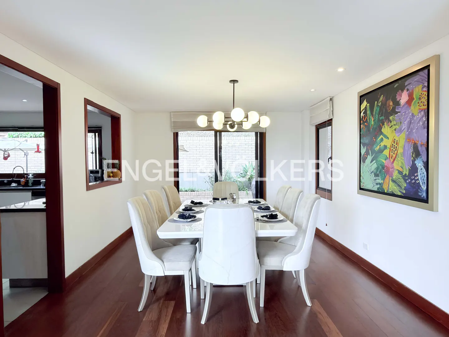 Modern dining room with white table, eight upholstered chairs, globe chandelier, and colorful abstract art on white walls.