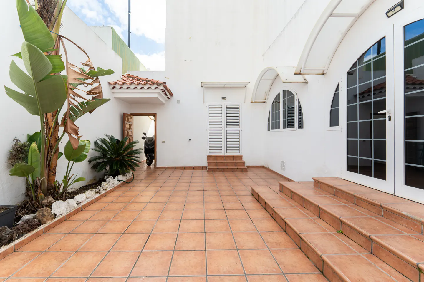 A tiled courtyard with white walls, plants, and a motorcycle parked inside an open doorway.