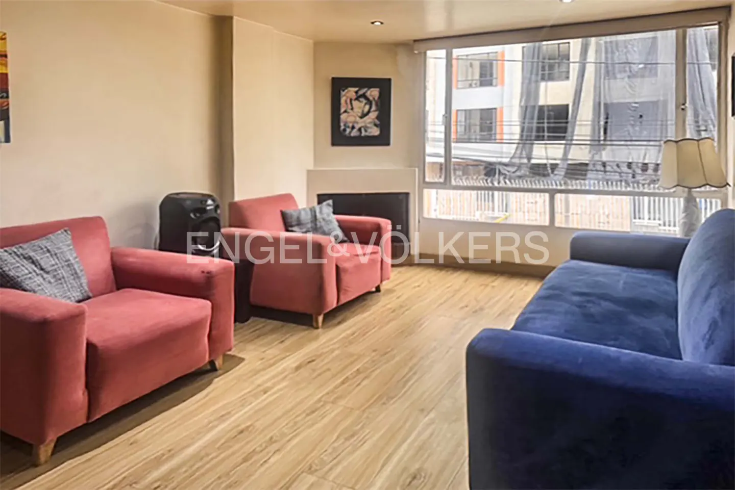 Living room with wood floors, red armchairs, and a blue sofa. A fireplace and large window are in the background.