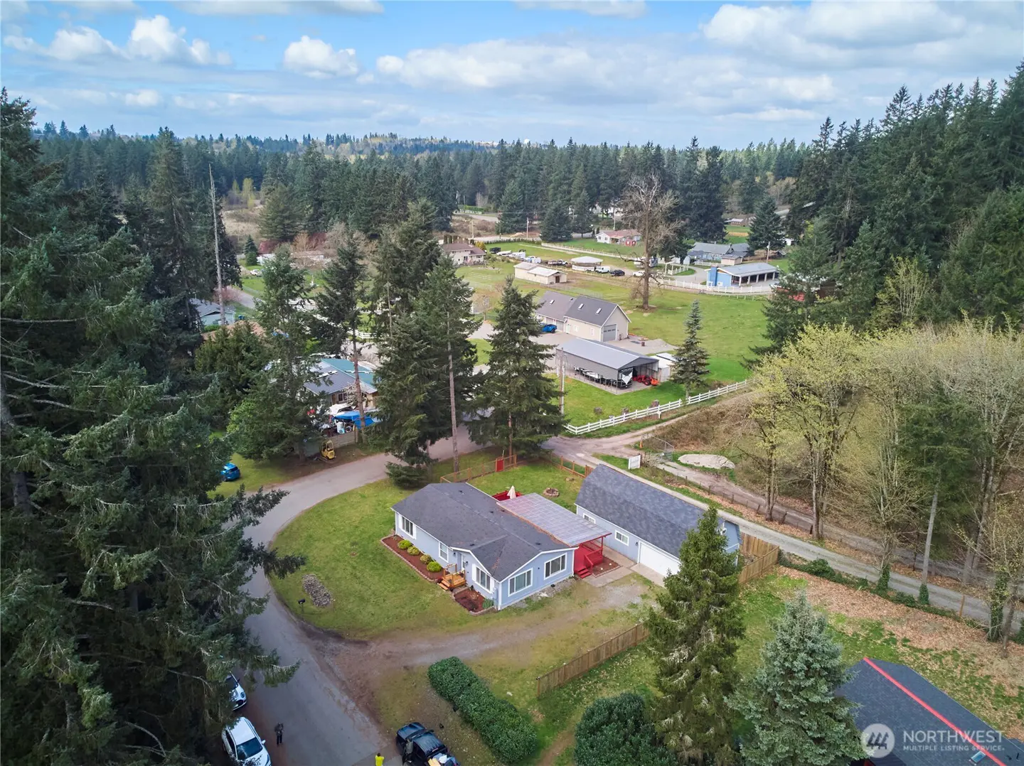 Aerial view of a light blue single-story house with a gray roof and a detached garage, surrounded by green lawns and tall trees.