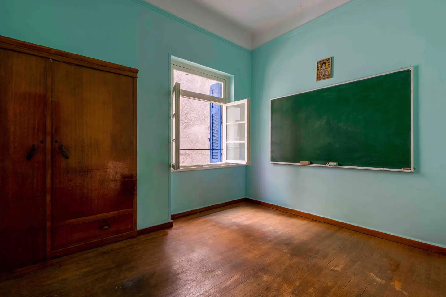 A light blue room with a wooden floor, wardrobe, open window, and a green chalkboard on the wall.