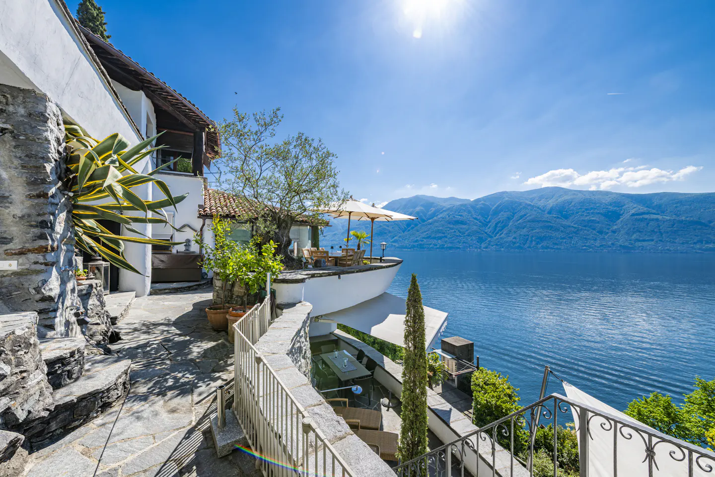 Stone path leads to a white villa with lake views. Outdoor seating areas with umbrellas overlook the blue water and mountains.