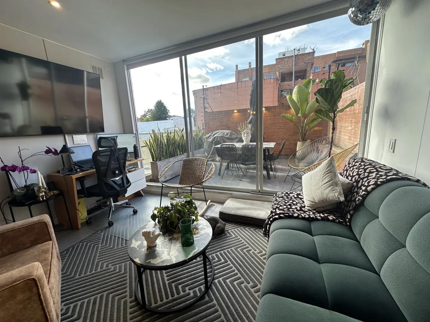 Modern living room with a teal sofa, patterned rug, home office area, and sliding doors opening to a furnished patio.