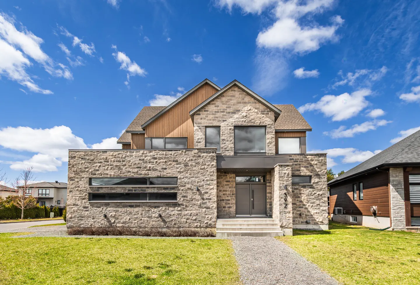 Modern two-story stone house with wood accents, double front doors, and a gravel path under a bright blue sky.