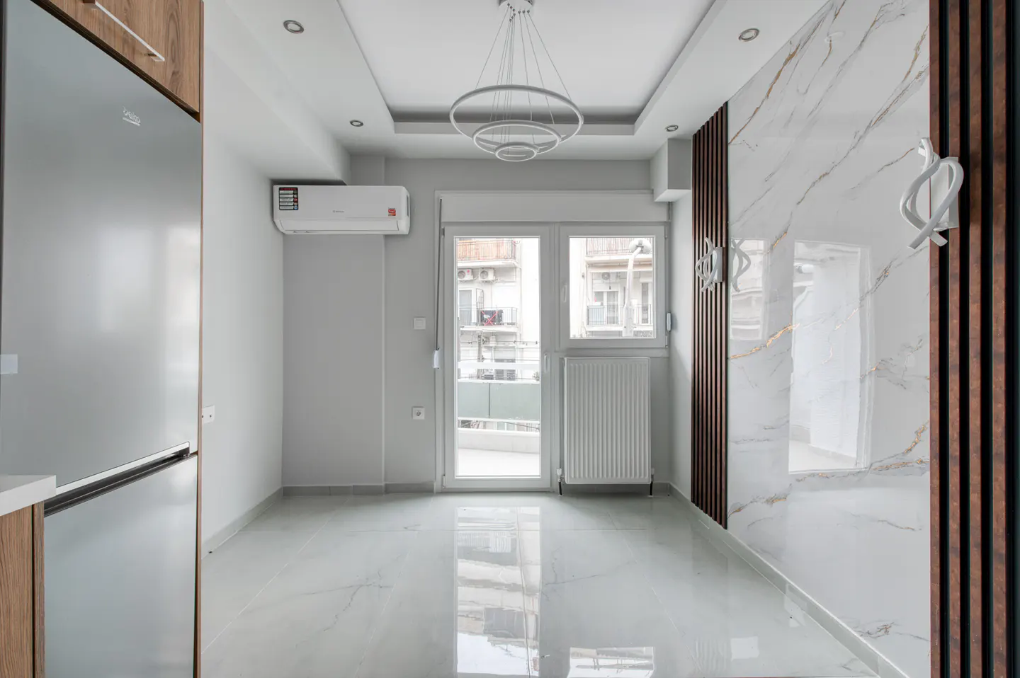 Bright, empty room with marble floors, a white radiator, and a modern chandelier. A silver refrigerator is on the left. A balcony is visible.