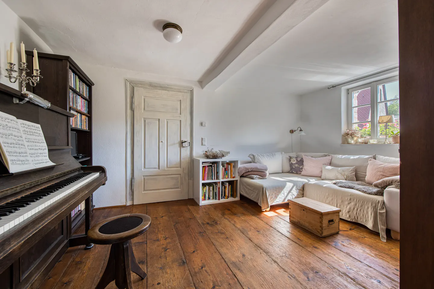 Cozy living room with a dark wood piano, stool, and bookshelf. A white couch sits near a window with plants. The floor is rustic wood.