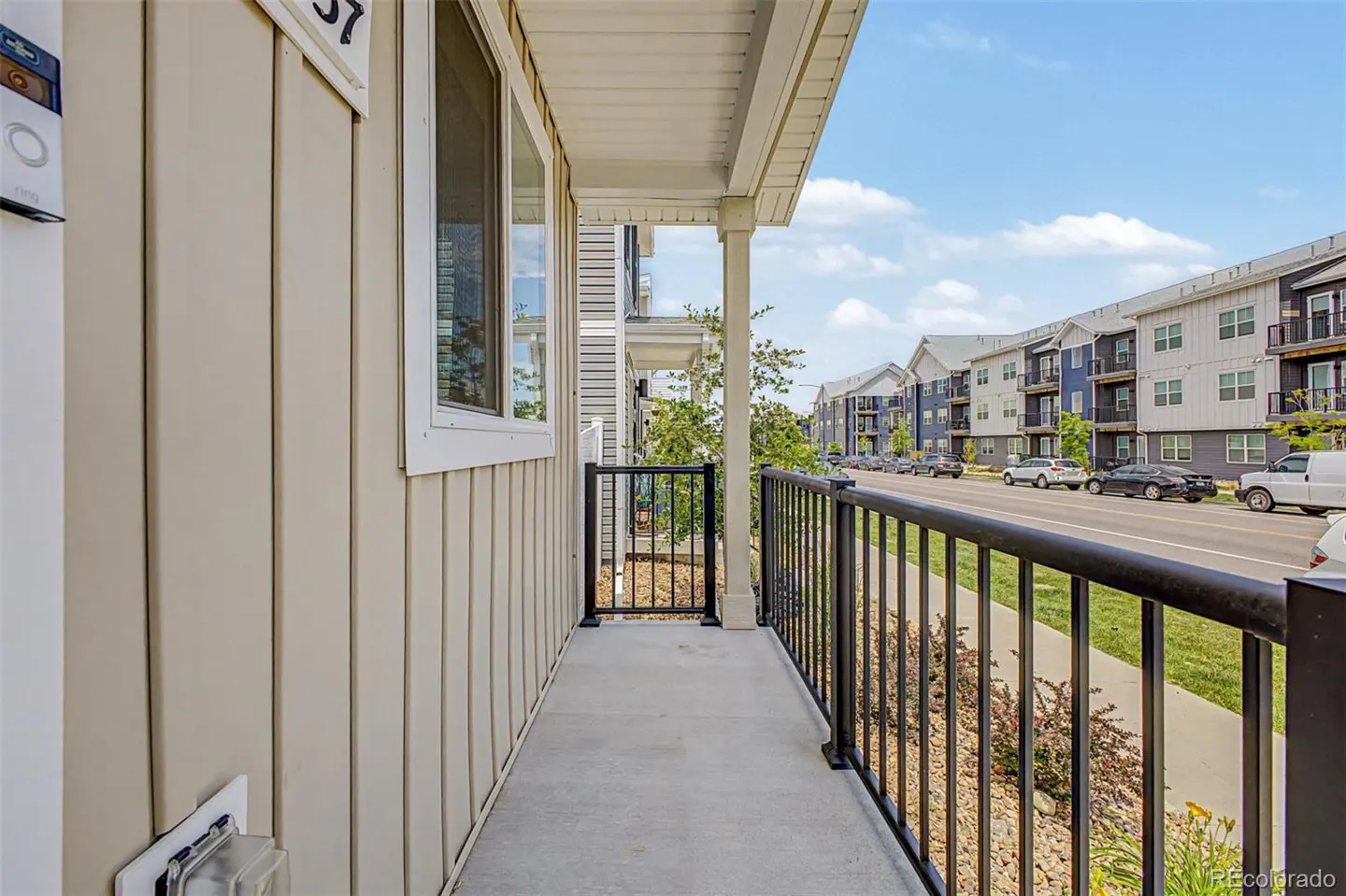 A view from a porch with black railings, looking out at a street with parked cars and apartment buildings. The building is beige with white trim.
