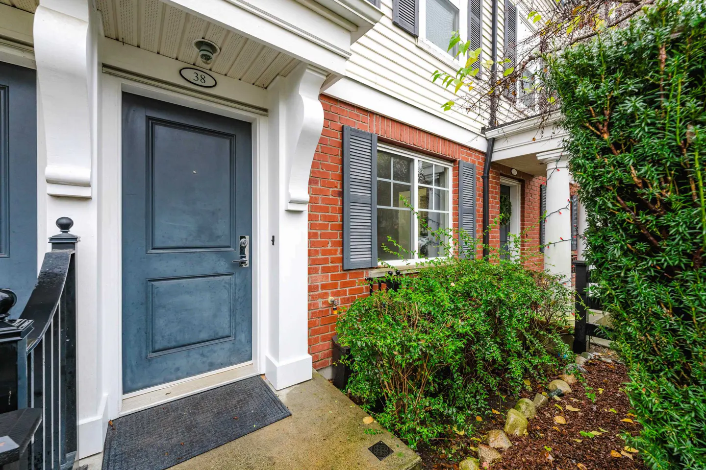 Front entrance of a townhouse with a blue door, red brick facade, white trim, and a small garden with a green bush.
