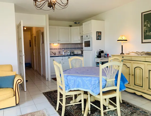 A bright apartment interior with a dining table, white kitchen cabinets, and a hallway in the background.
