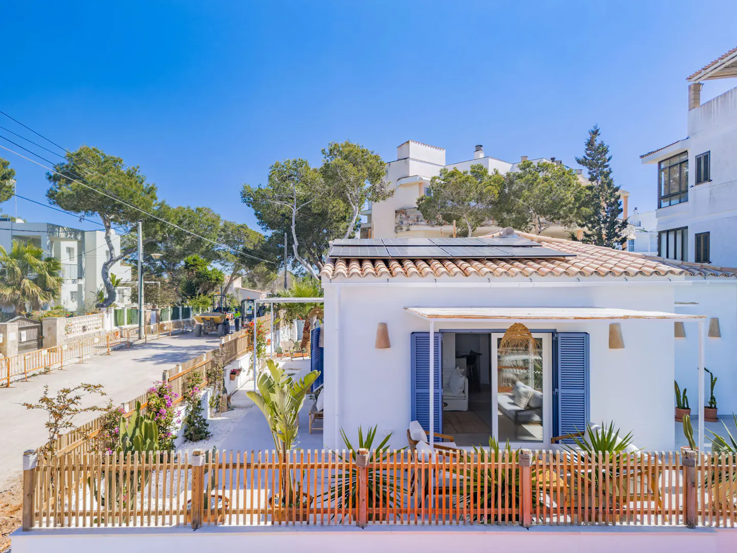 Exterior view of a white house with blue shutters, a tiled roof with solar panels, and a wooden fence. Palm trees and other greenery surround the house.