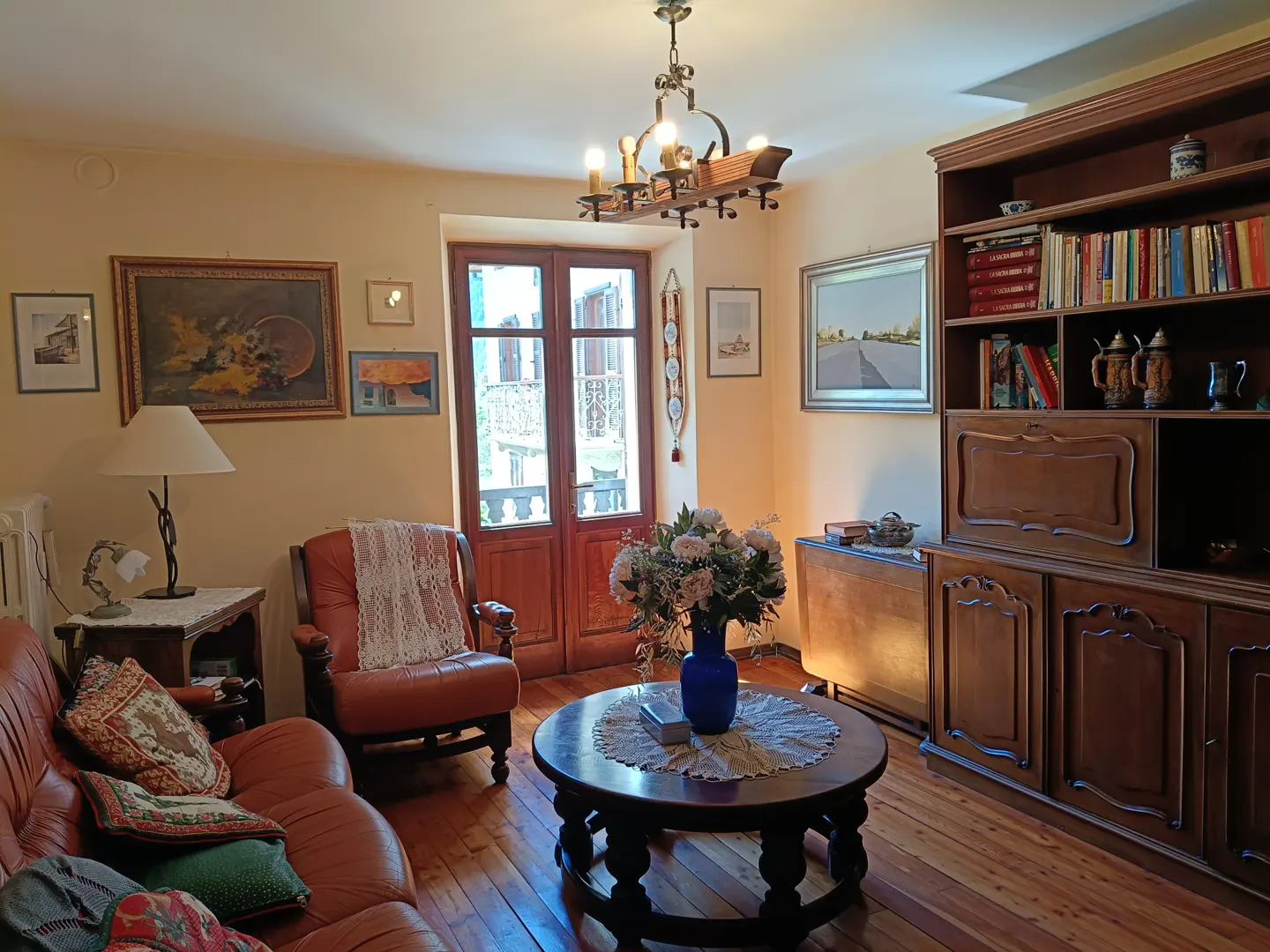 Living room with wood floors, a leather sofa, a round coffee table with flowers, and a wooden bookcase. Paintings adorn the walls.