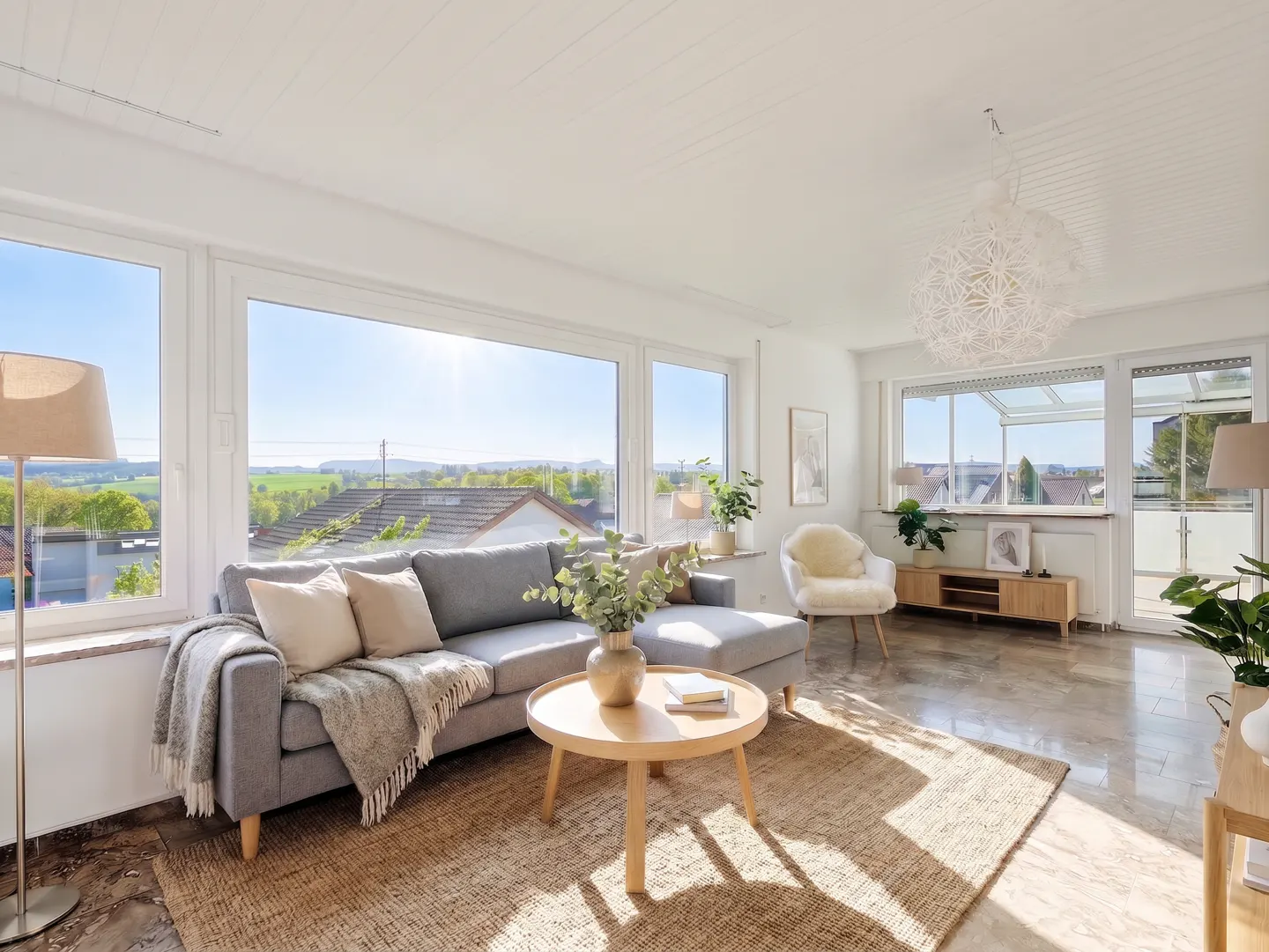 Bright, airy living room with a gray sofa, round wood table, and large windows overlooking a green landscape. A jute rug adds warmth to the marble floor.
