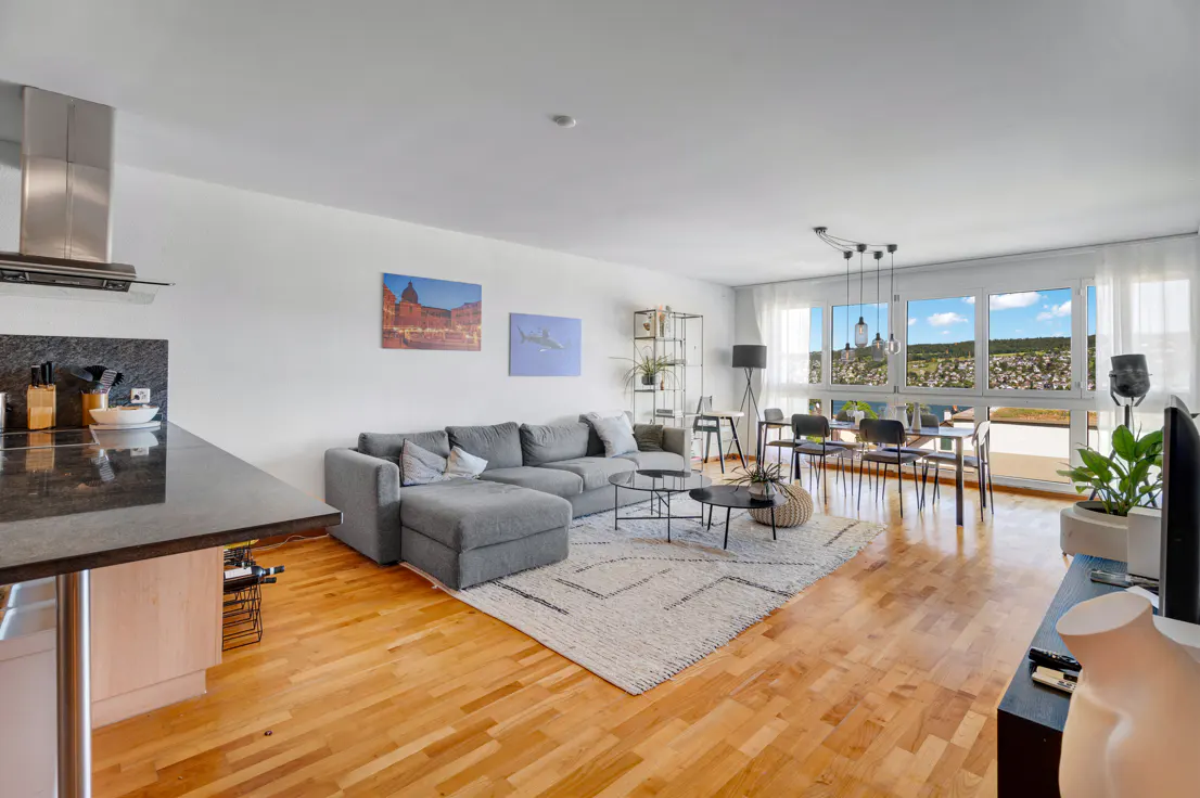 Open-plan living area with a grey sectional sofa, dining set, hardwood floors, and large windows with a scenic view.