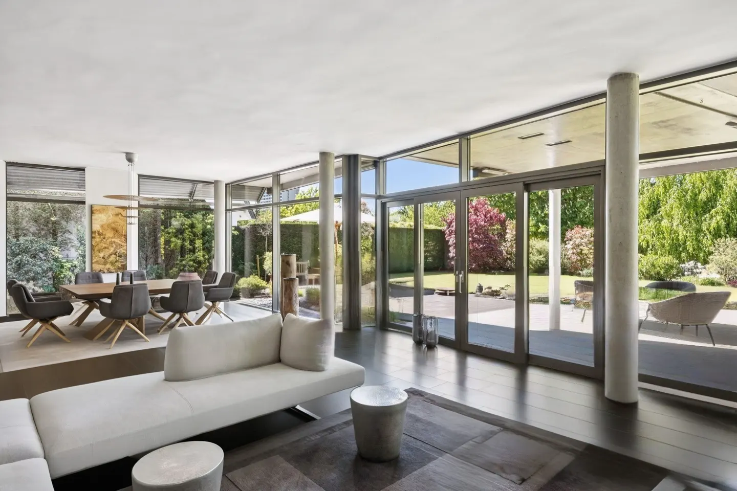 Bright, modern living room with a white sofa, dark wood floors, and floor-to-ceiling windows overlooking a lush green garden. Dining area visible.