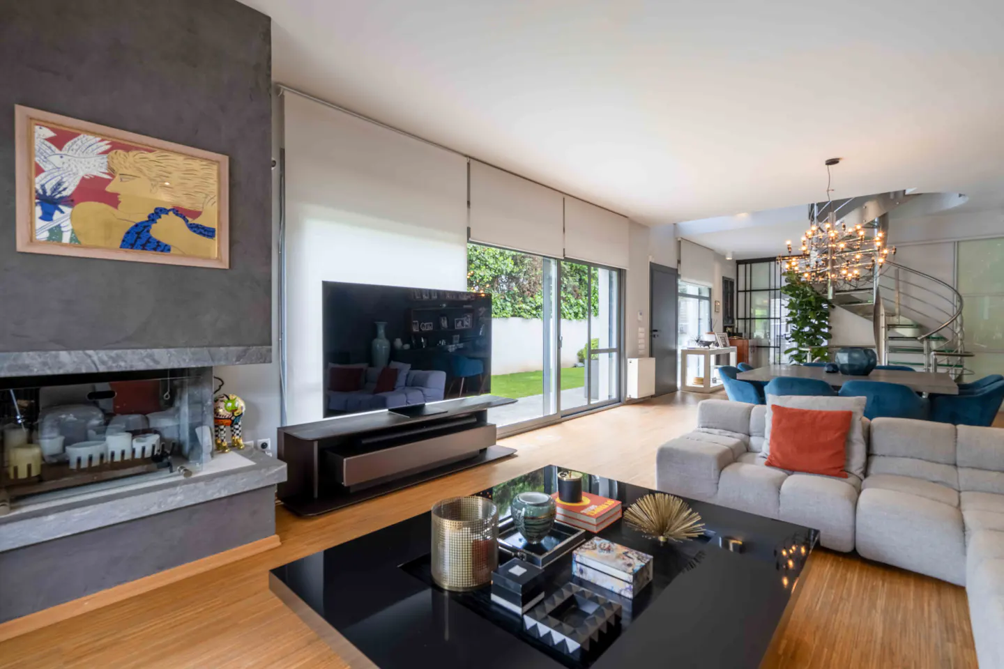 Bright living room with a gray sofa, black coffee table, and a large TV. A spiral staircase is visible in the background.