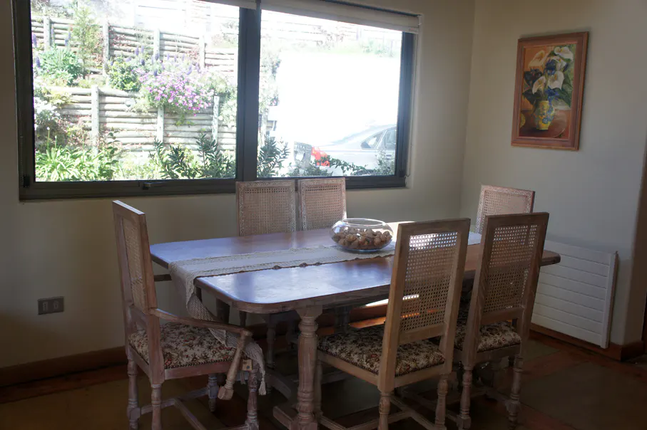 Dining room with a wooden table, six chairs, and a bowl of fruit. A window shows a garden and a painting hangs on the wall.