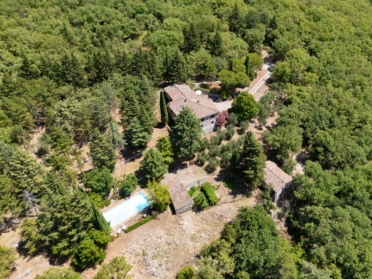 Aerial view of a stone house with a red tile roof, a pool, and a dirt road, surrounded by a dense green forest.
