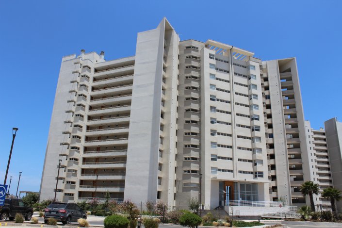 Exterior view of a tall, white apartment building with balconies under a clear blue sky. Cars are parked in front.
