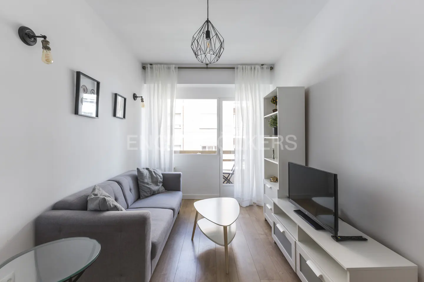 Bright living room with gray sofa, white walls, wood floors, and balcony access. Modern light fixture hangs from the ceiling.