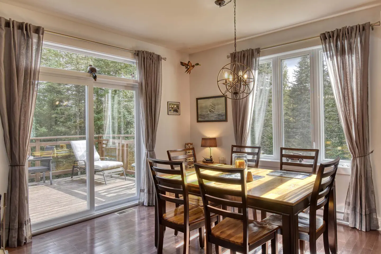 Dining room with wood table, chairs, and hardwood floors. Large windows with gray curtains overlook a deck and forest. A spherical chandelier hangs above.