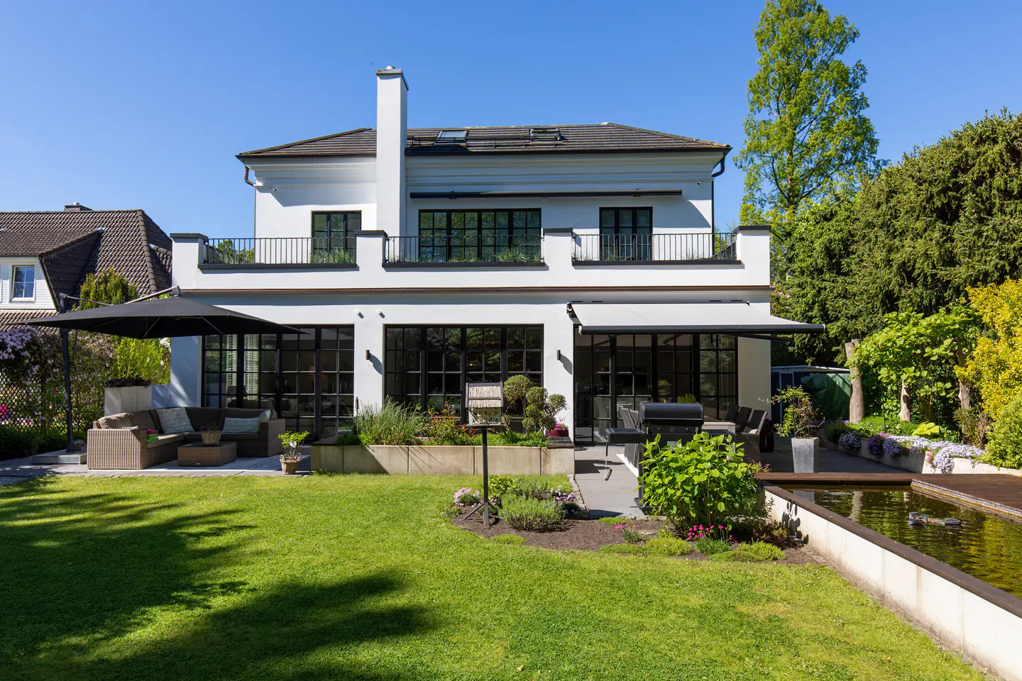 Back view of a white two-story house with black framed windows, a green lawn, and a pond.