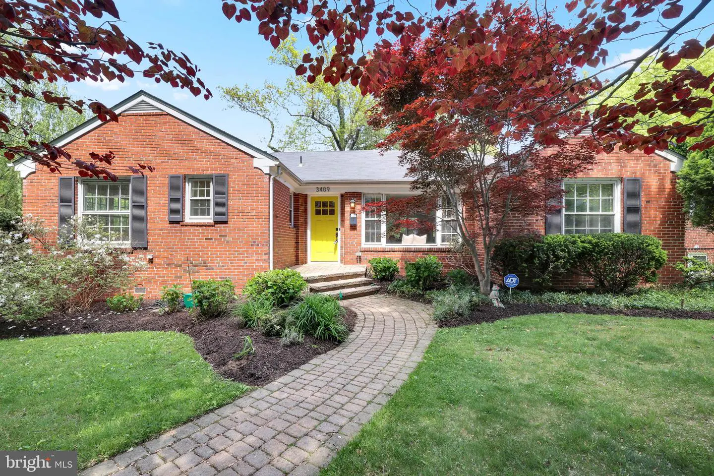 Red brick house with a bright yellow front door and a curved brick walkway leading to the entrance. Green lawn and trees surround the house.