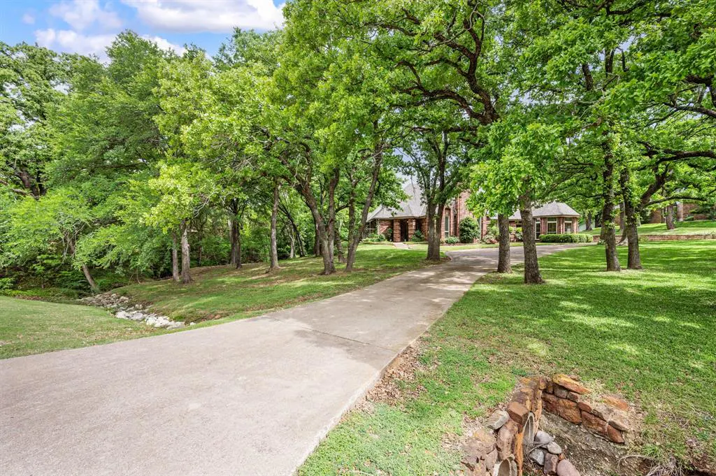 Long driveway leading to a brick house, surrounded by green trees and grass. Blue sky with white clouds.