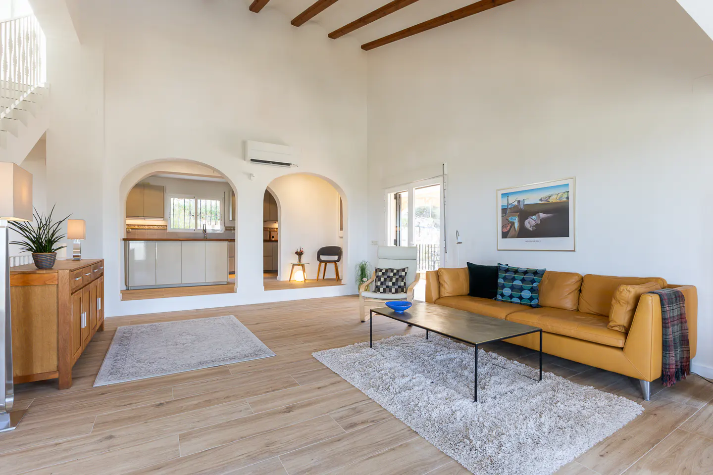 Bright living room with white walls, wood floors, and exposed beams. A tan leather sofa sits on a white rug. Arched doorways lead to the kitchen.