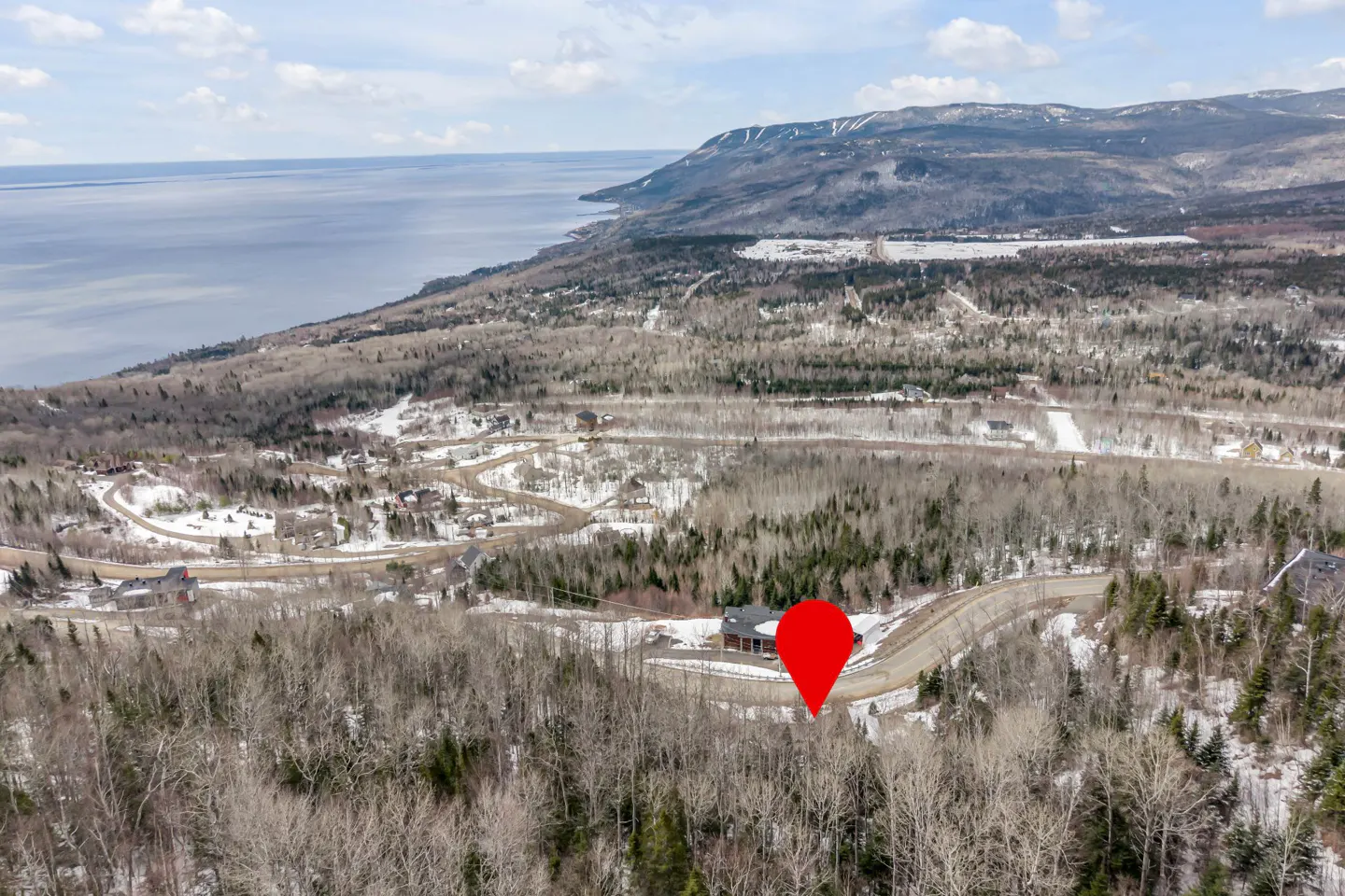 Aerial view of a house with a red location pin, surrounded by trees, with a lake and mountains in the background.