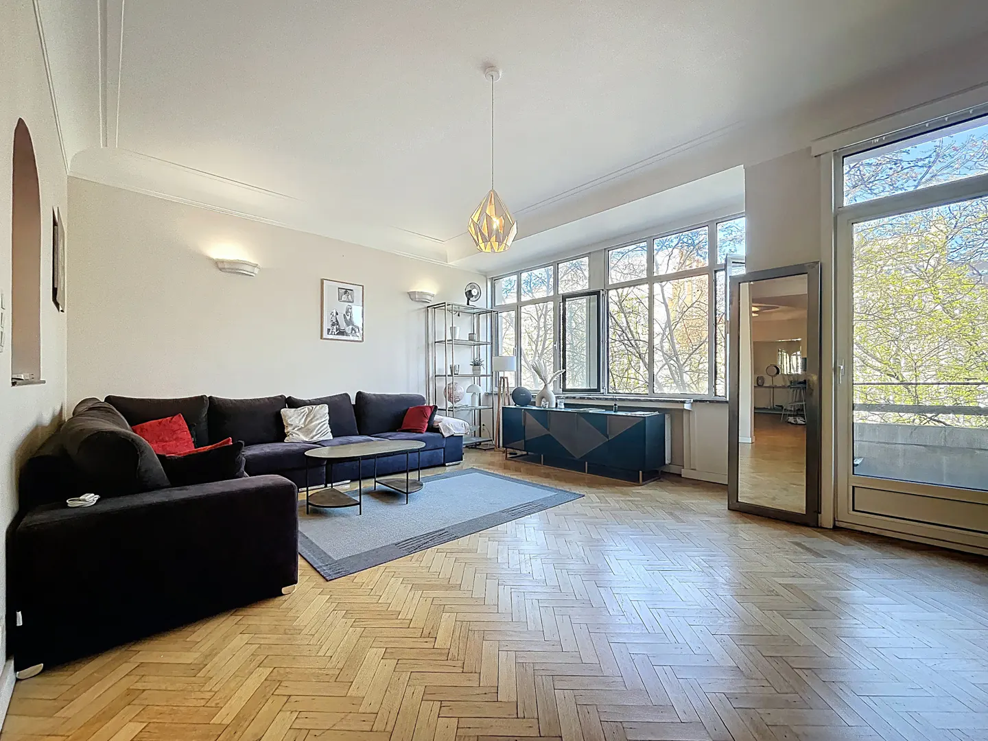 Bright living room with herringbone wood floors, large windows, and a dark blue sectional sofa with red pillows.