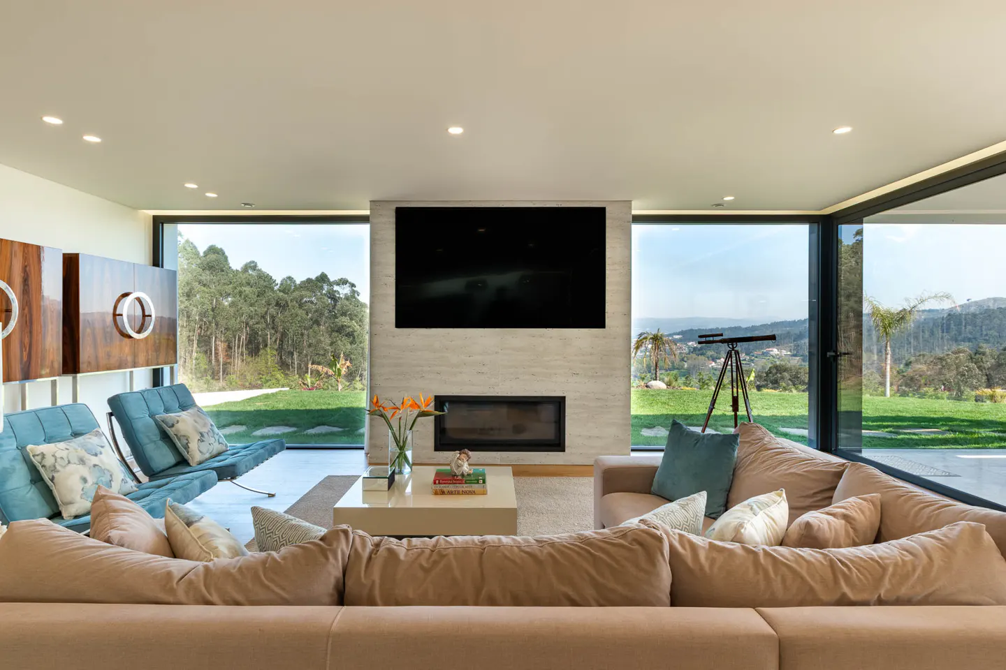 Bright living room with beige sofa, blue chairs, and large windows overlooking a green lawn and trees. A TV and fireplace are centered on the wall.