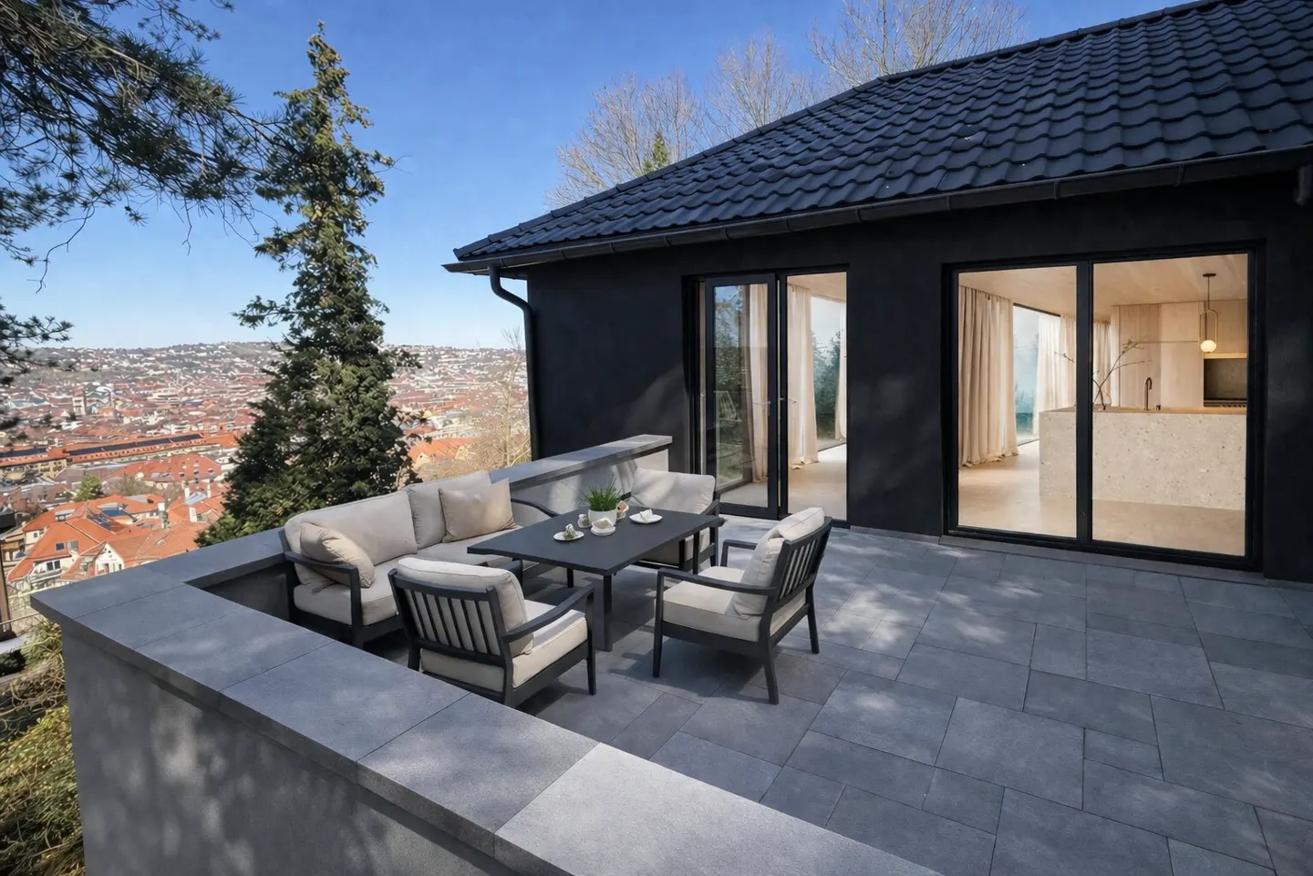 Modern black house with a stone patio, outdoor seating, and a panoramic view of a city below under a clear blue sky.