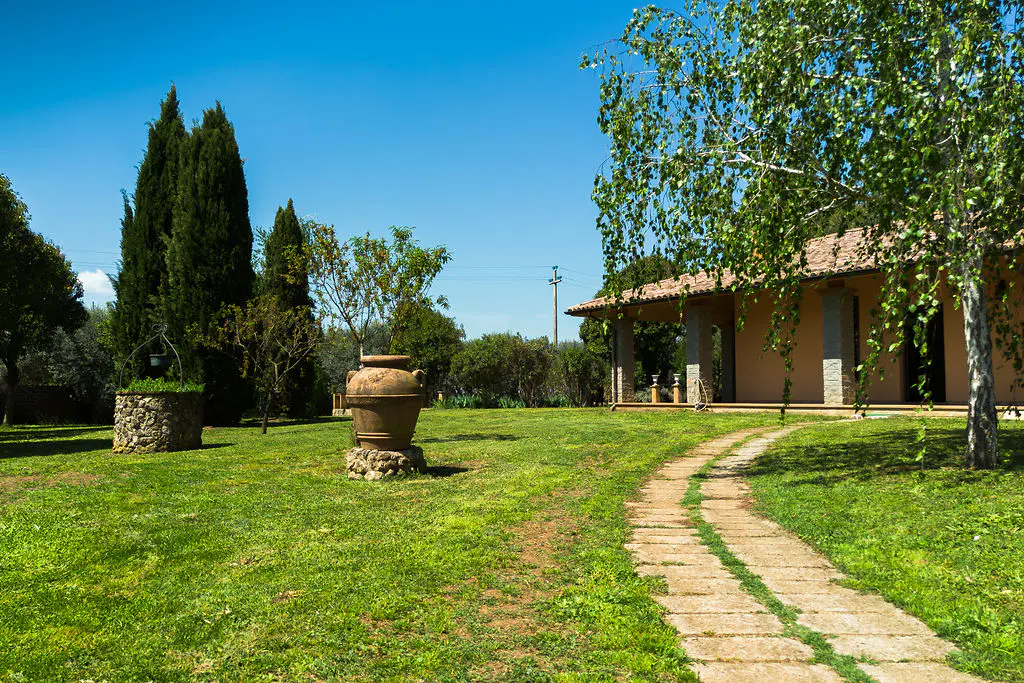 Lush green lawn with stone path leading to a peach-colored house with a tiled roof. A large clay pot sits on a stone base.