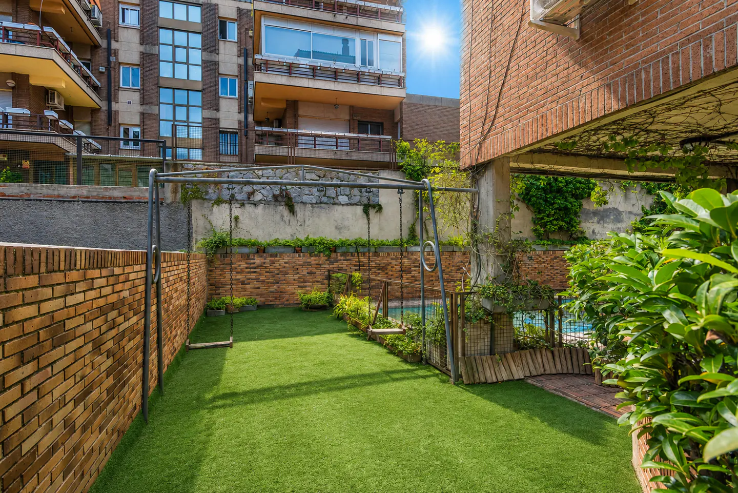 A backyard with artificial grass, a metal swing set, brick walls, and a small pool area under a sunny sky.