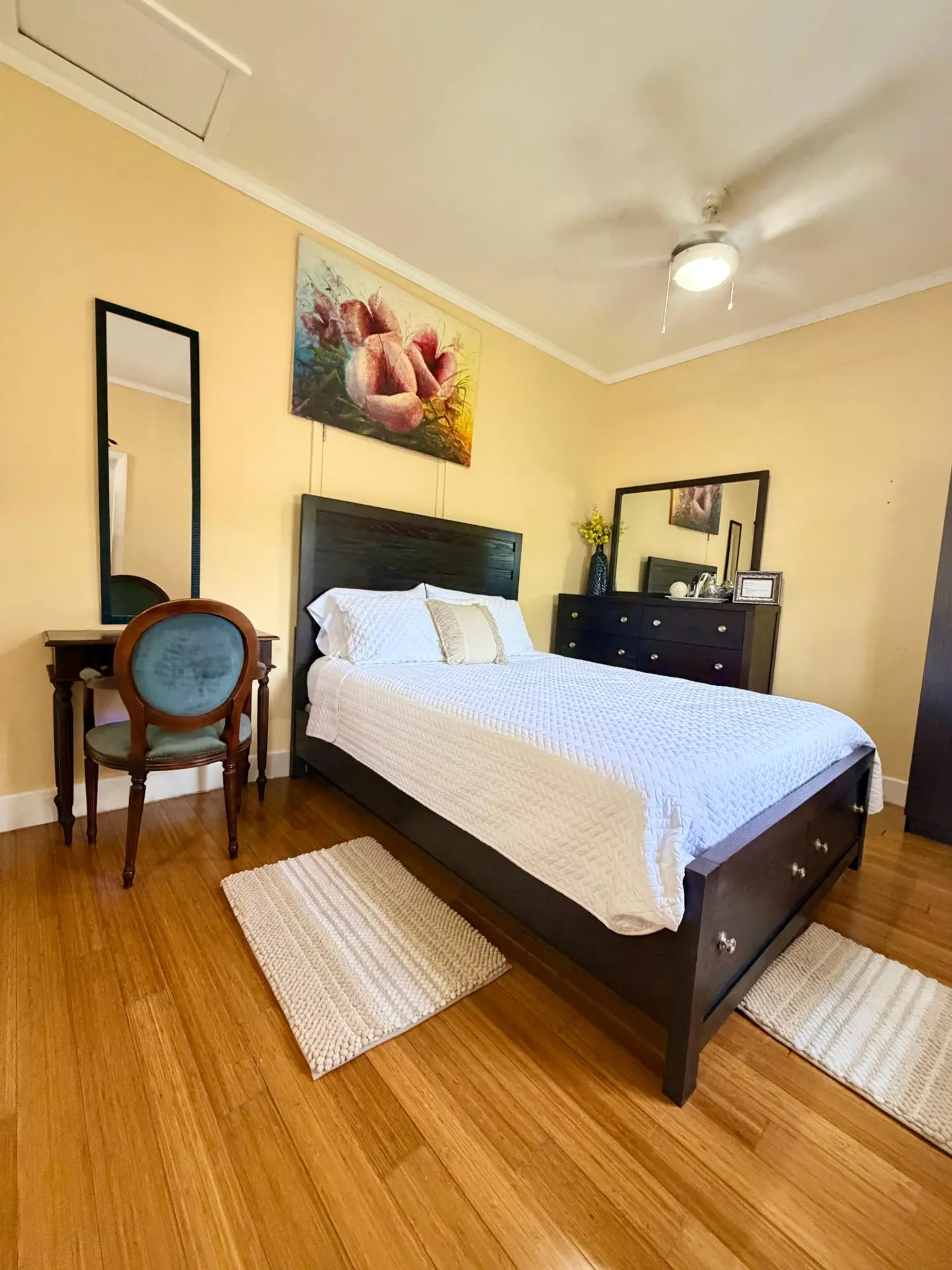 Bedroom with a dark wood bed, white bedding, and a floral painting above the bed. A desk and chair are to the left.