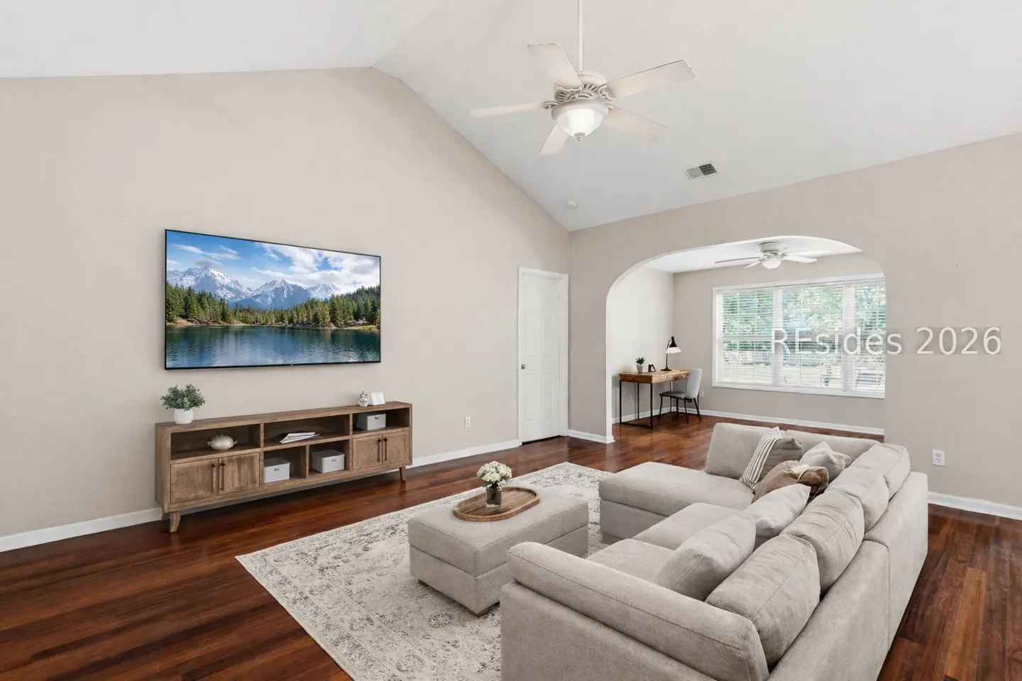 Living room with vaulted ceiling, ceiling fan, TV with mountain scene, gray sectional sofa, and wood floors.