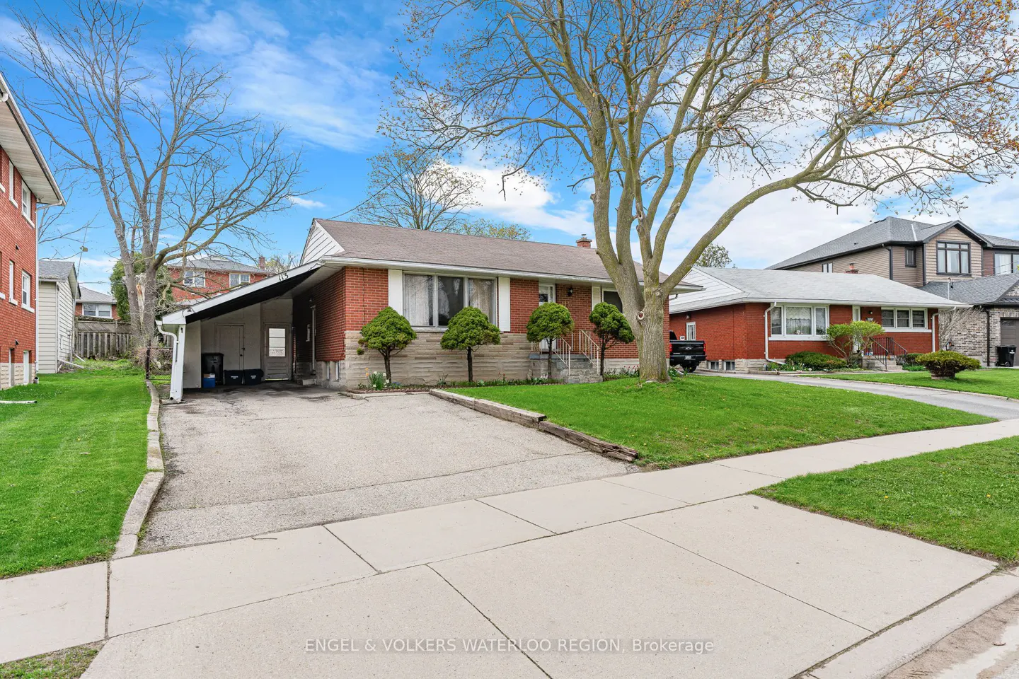 A red brick bungalow with a carport, a concrete driveway, and a green lawn on a sunny day.