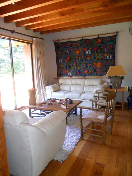 Living room with wood floors, exposed beams, and white sofas. A colorful textile hangs above the sofa. A wooden coffee table sits on a white rug.